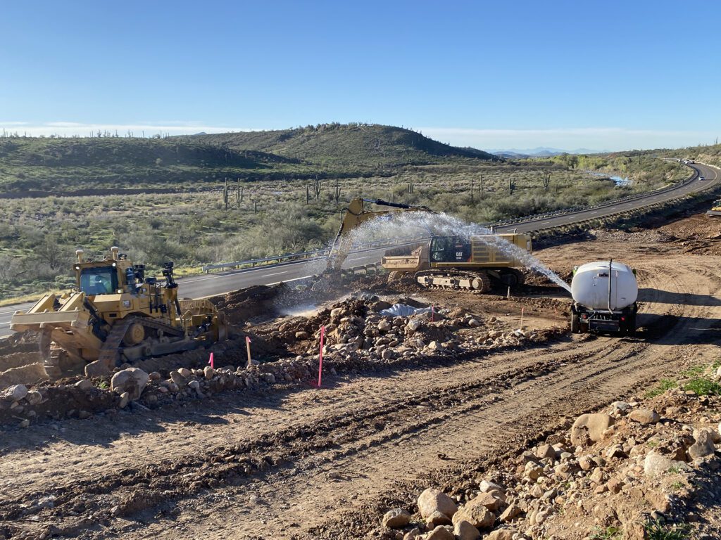 Bulldozer at a road construction site moving earth while a water truck sprays to control dust, showcasing Civil Engineering News and Events.