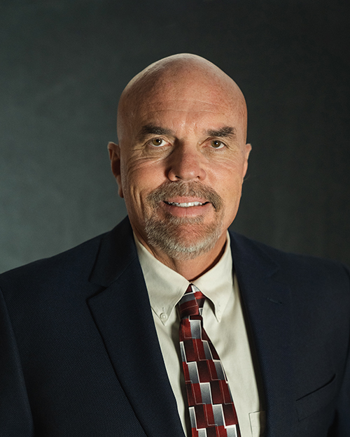 A bald man from the QT Executive Team wearing a suit with a patterned tie smiling at the camera against a dark background.