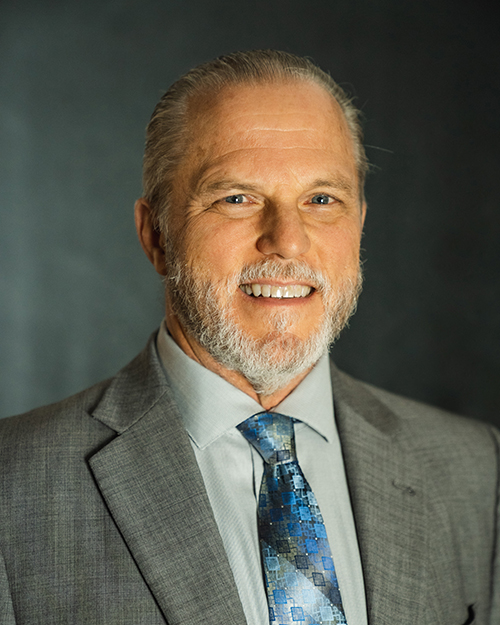 Portrait of a smiling man with a beard in a suit and tie from the QT Executive Team.