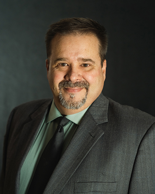 A professional portrait of a smiling man from the QT Executive Team, dressed in a suit and tie against a dark background.