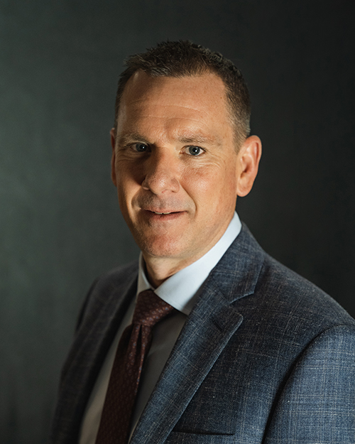 A professional man in a suit, from the QT Executive Team, posing for a formal portrait against a dark background.