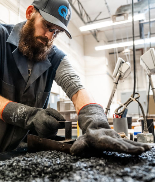 A bearded worker from Quality Testing LLC in a cap and safety gloves precision-measuring a metal part in an industrial environment.