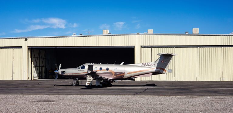 A small private airplane parked outside a civil engineering services hangar under a clear blue sky.