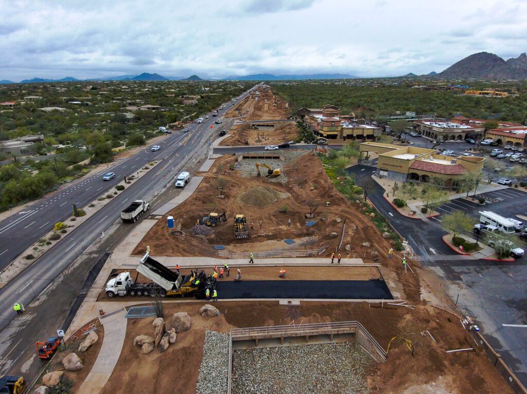 Aerial view of a road under construction with equipment and workers, providing civil engineering services, showing partial lane closures and adjacent commercial buildings.