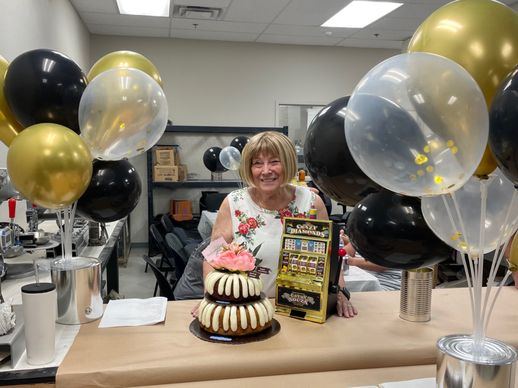 A smiling Cathy Souza celebrates her retirement with a cake, flowers, balloons, and an award in a decorated room.