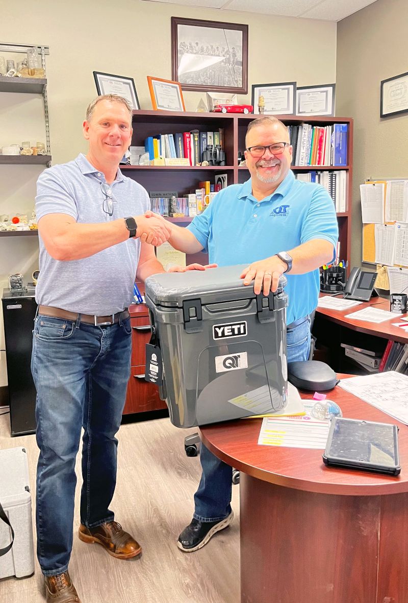 Two men shaking hands in an office setting, with one handing over a yeti cooler as Jaye Richardson celebrates 5 years at QT.