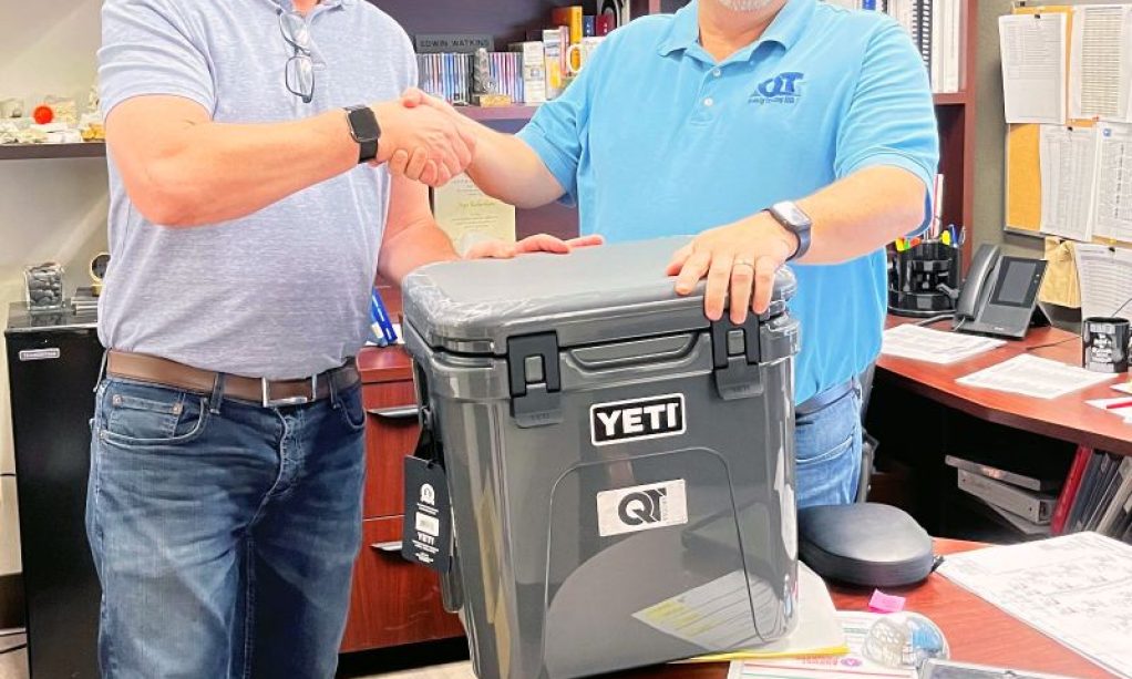 Two men shaking hands in an office setting, with one handing over a yeti cooler as Jaye Richardson celebrates 5 years at QT.